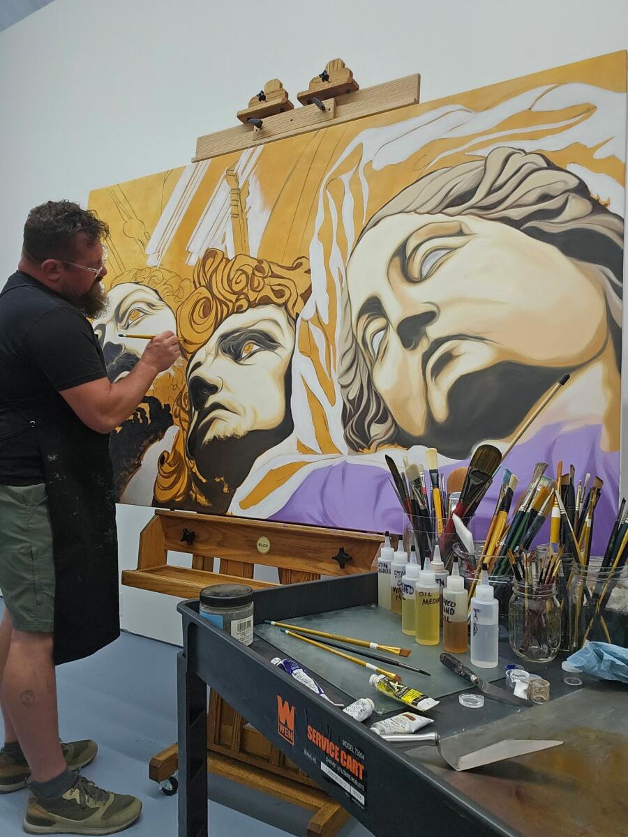 Josh Niccolai-Belfi at work in his studio, painting a large canvas of classical figures in gold and violet, brushes and bottles on the bench in front of him